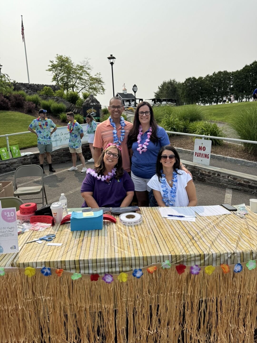 photo of a group of four individuals at the registration table. all are wearing leis, smiling and are sitting at a table with a grass skirt around the edge of the table