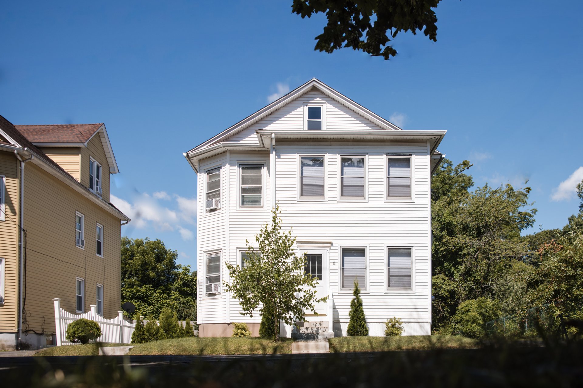 Two-story white house with multiple windows, surrounded by greenery and a neighboring yellow house under a clear blue sky.