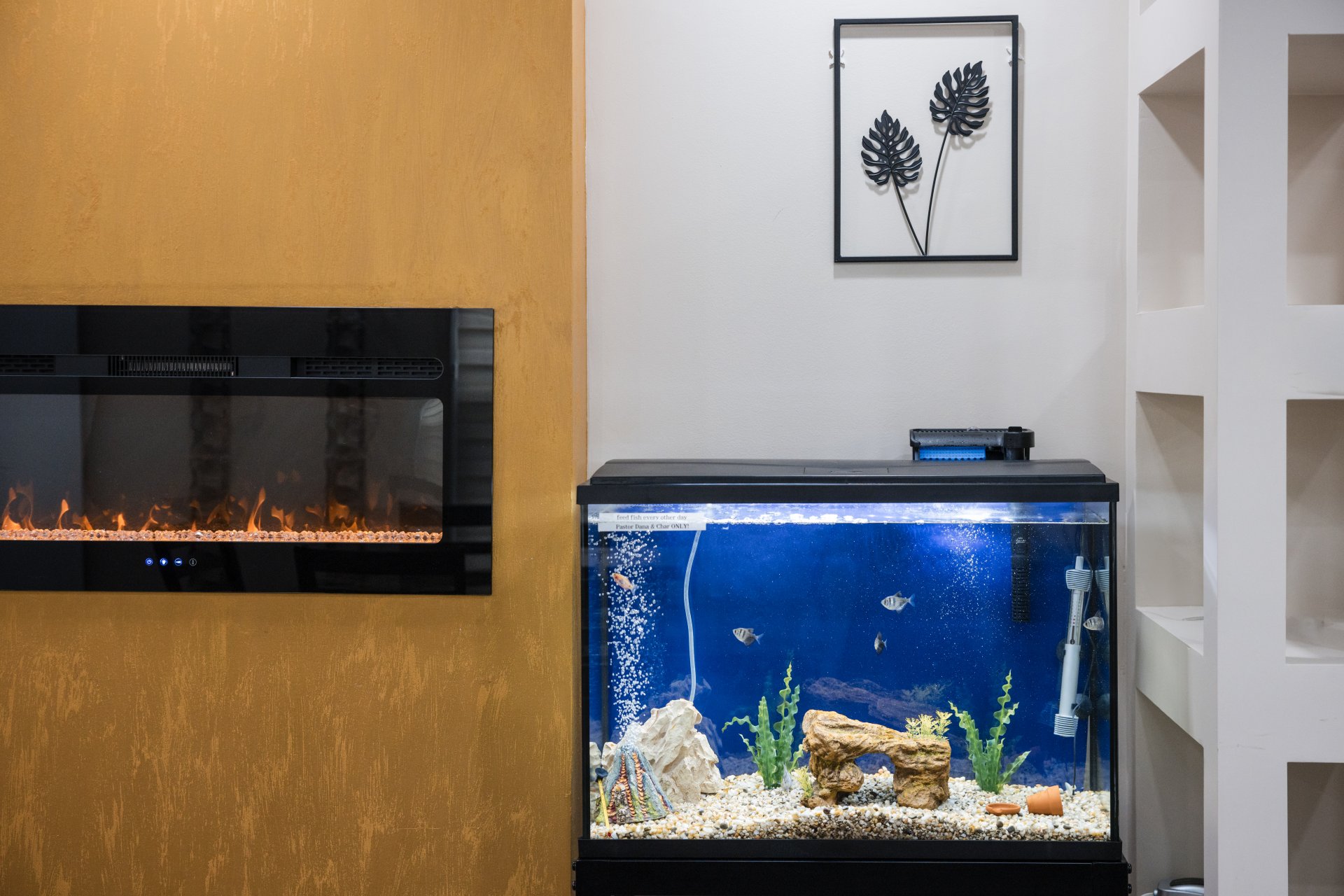An aquarium with fish and aquatic plants sits next to an electric fireplace on a beige wall. A framed leaf decor hangs above.