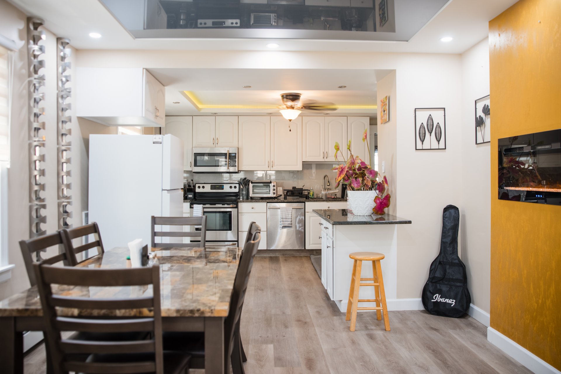 Open-plan kitchen and dining area with white cabinets, stainless steel appliances, wooden table and chairs, a guitar case, and decorative flowers.