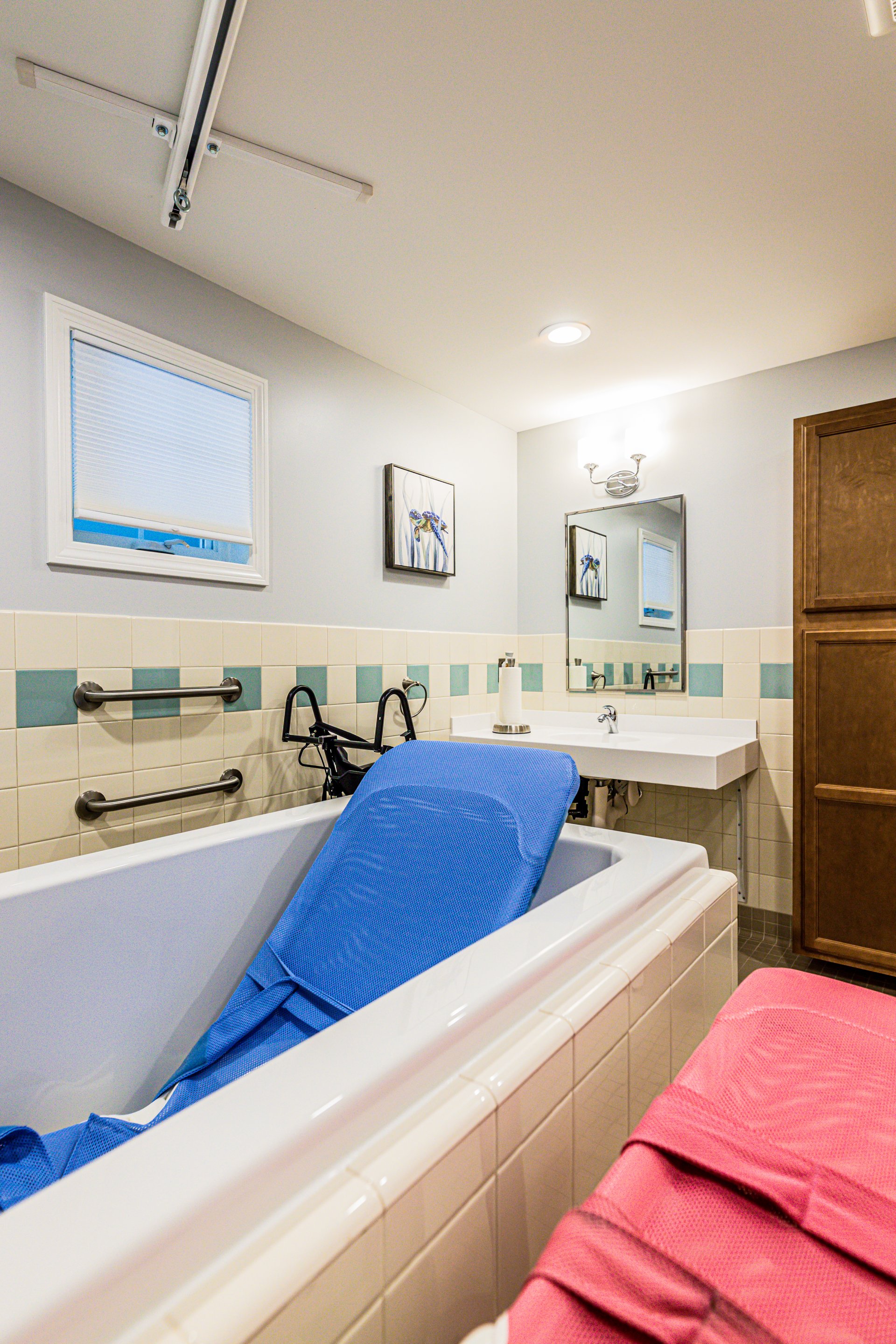 A bathroom with an accessible bathtub featuring a blue transfer bench, grab bars, a rollator walker, a sink, a mirror, and wall art on light-colored tiled walls.