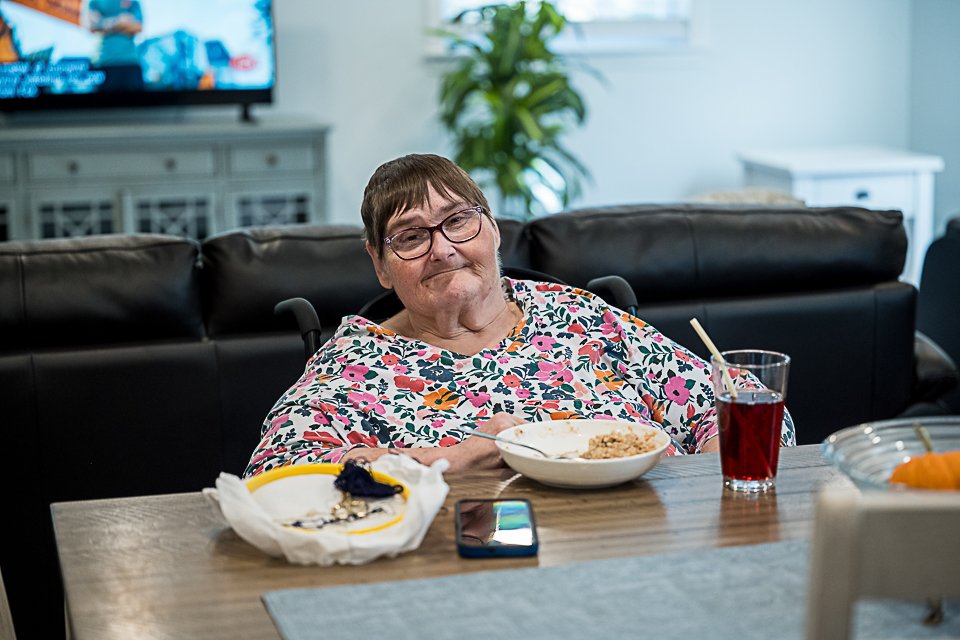 Photo of Michele sitting at the table with her embroidery, food, drink & cell phone in front of her