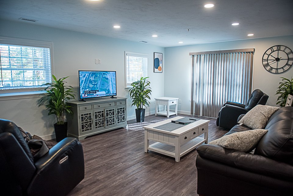 Photo of a well-lit living room with couches, coffee table, tv & tv stand with two plants on either side of them.