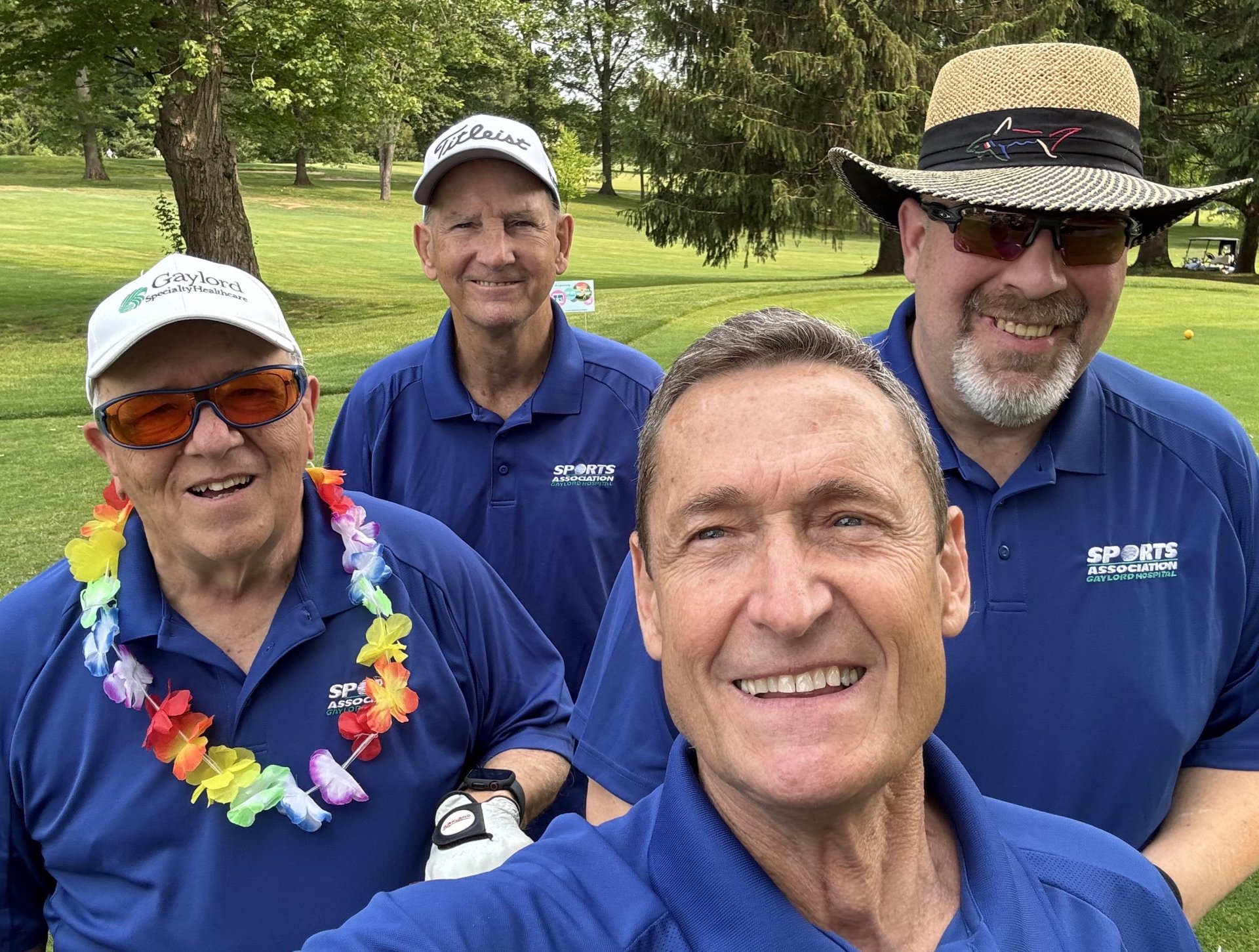 Group photo of four individuals in blue golf polos, some with hats, some with sunglasses and a tropical lei on, smiling on the golf course