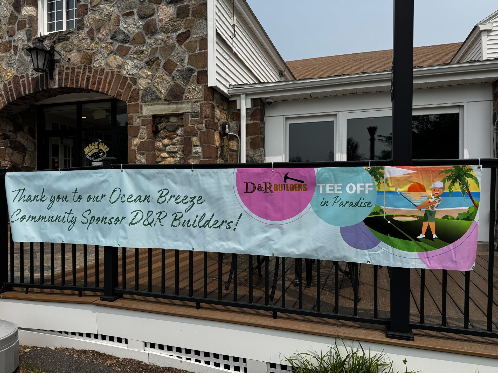 Image of a large bright banner hanging on a railing. Banner says Thank you to our ocean breeze community sponsor D&R Builders! with a graphic in the top right corner including the company's logo and a tropical background with a golfer teeing off