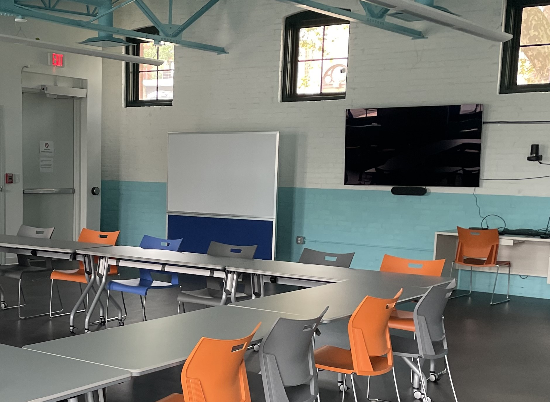 Image of conference room filled with tables & colorful chairs. A rollable whiteboard  & large tv are also shown in the conference room